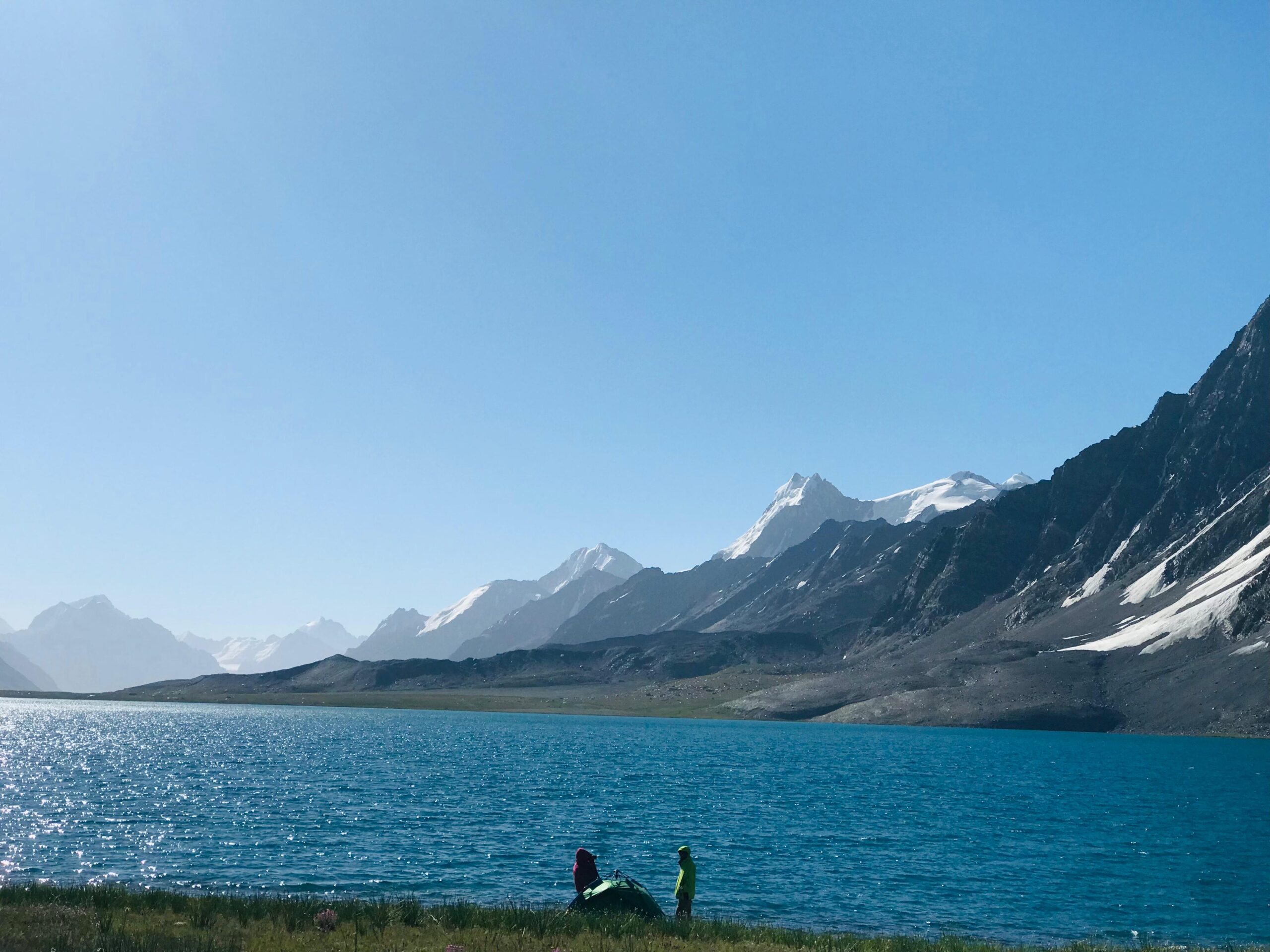 A trekking group stands near the crystal-clear shoreline of Karambar Lake with snowy peaks in the background under clear blue skies.