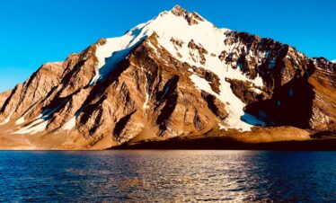 Still waters of Karambar Lake reflecting snow-covered mountain peaks under a pastel-colored sky.