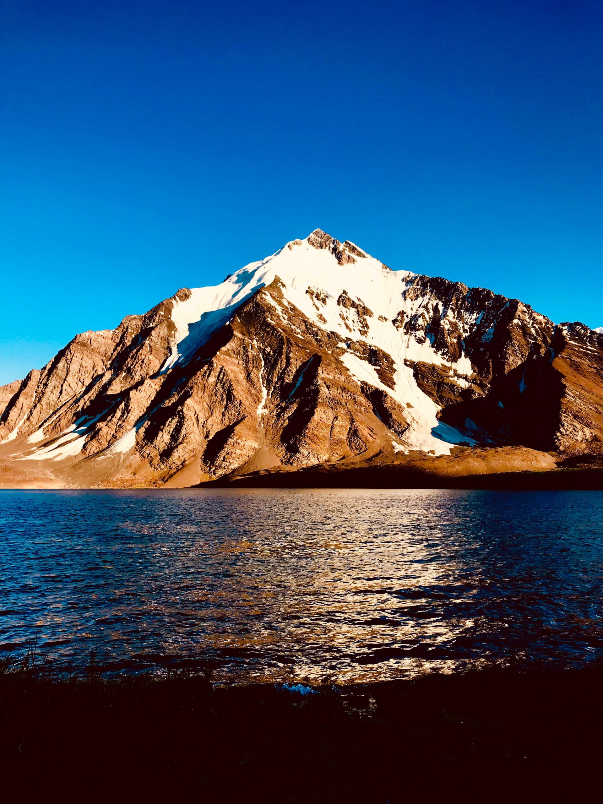 Still waters of Karambar Lake reflecting snow-covered mountain peaks under a pastel-colored sky.